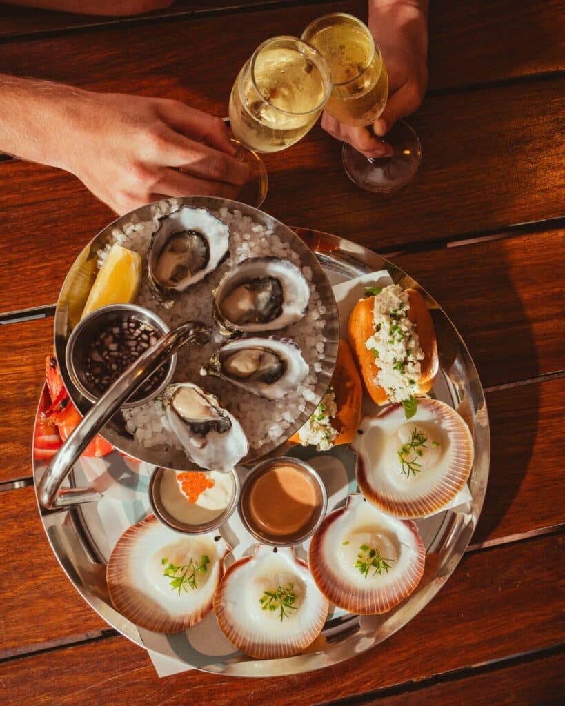 Um prato de frutos do mar com ostras, vieiras e pão, servidos no gelo com molhos, em uma mesa de madeira. Duas mãos seguram taças de champanhe. Está no Opera Bar em Sydney.