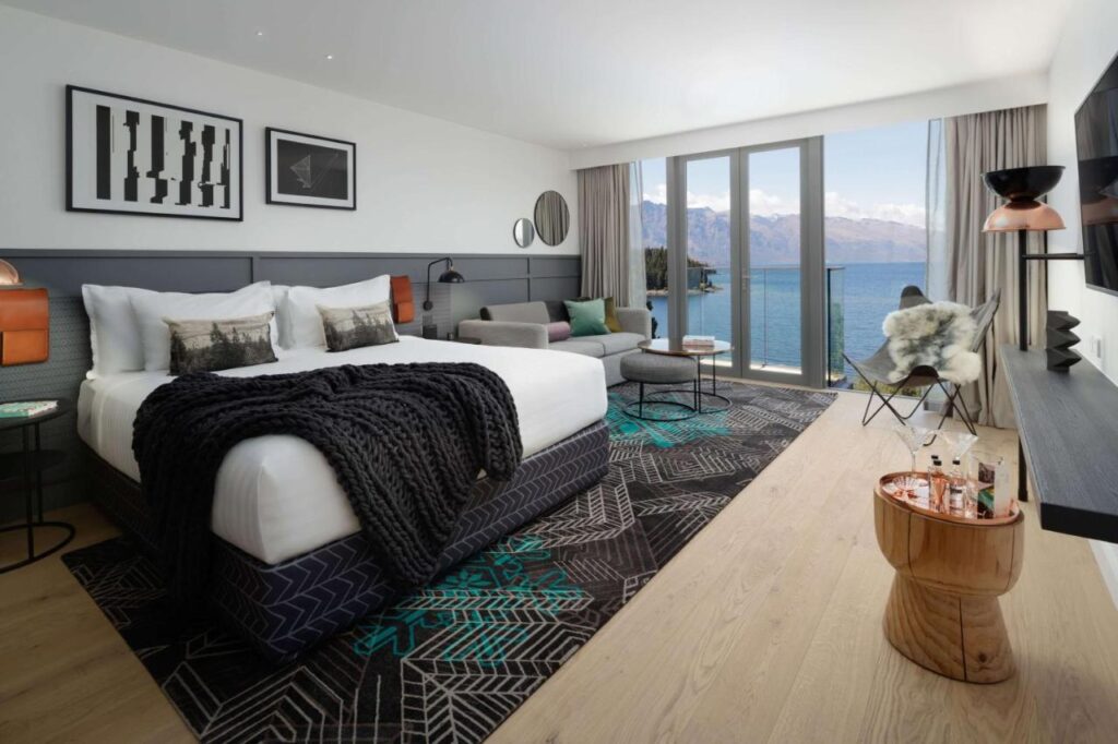 Quarto do hotel QT Queenstown com uma cama grande, vista para a montanha e o lago através de portas de vidro, área de estar e decoração elegante.