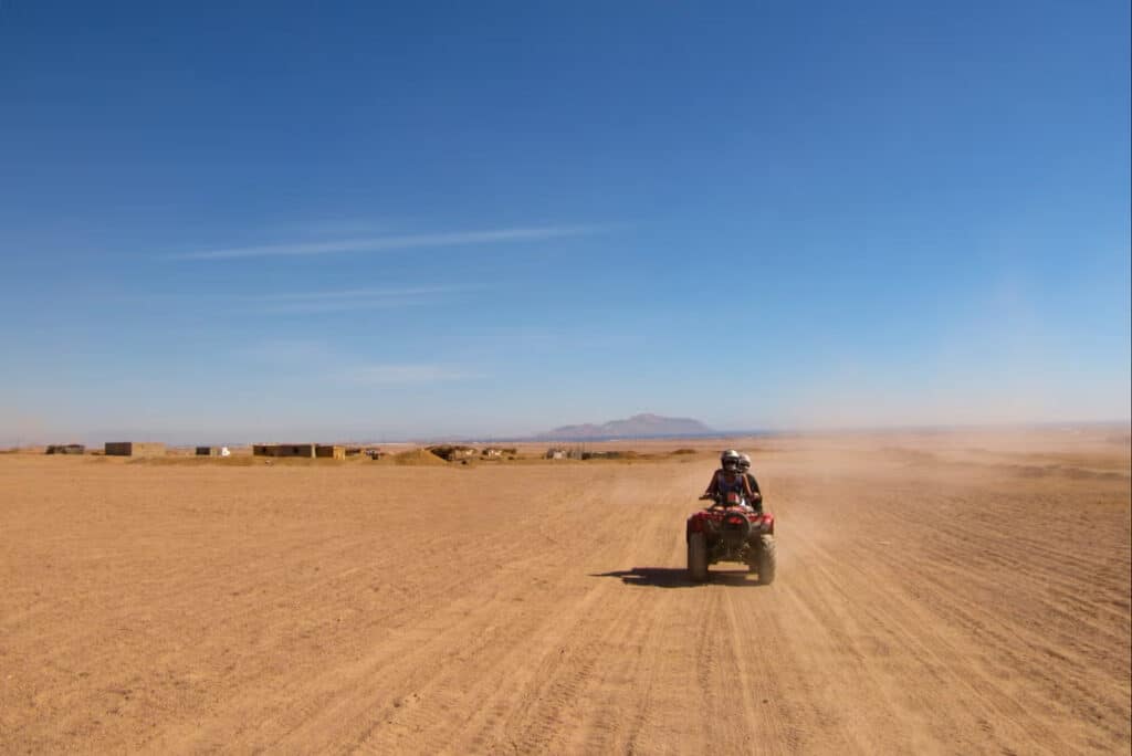 Uma pessoa em um quadriciclo por Agadir atravessa uma vasta paisagem desértica sob um céu azul claro com montanhas distantes no horizonte. Imagem para ilustrar post sobre o que fazer no Marrocos.