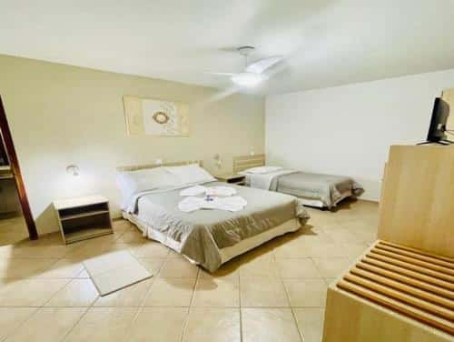 O quarto do Ózera Hotel Fazenda, no Paraná é espaçoso e decorado de maneira simples e funcional. Ele possui uma cama de casal e uma cama de solteiro, ambas com roupas de cama em tons neutros de cinza e branco. No centro da cama de casal, há toalhas dobradas em forma de cisne, trazendo um toque de acolhimento e carinho na recepção. O ambiente tem piso de cerâmica em tom claro, e a iluminação suave realça a tranquilidade do espaço. A mobília é minimalista, com uma cômoda de madeira clara e um ventilador de teto que complementa o conforto do quarto.