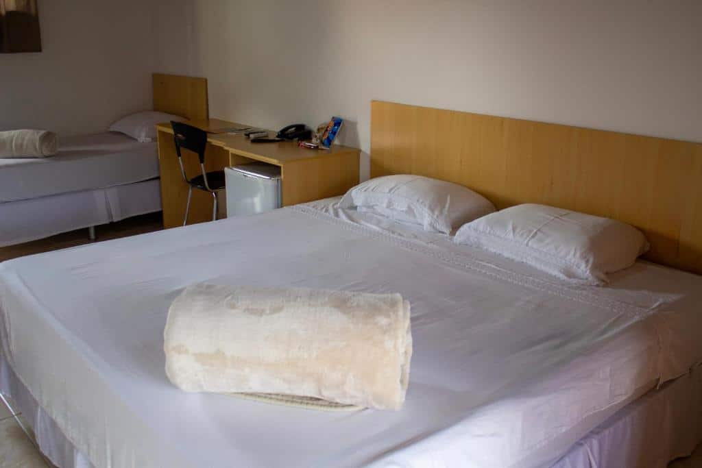 O quarto do GoldMen Hotel Fazenda Guarapuava, no Paraná, apresenta uma decoração simples e confortável. Ele possui uma cama de casal grande com lençóis brancos e uma manta dobrada sobre ela. Ao lado, há uma cama de solteiro adicional, sugerindo que o espaço pode acomodar mais de uma pessoa. O quarto é equipado com uma mesa de trabalho de madeira clara, sobre a qual há um telefone, objetos de uso pessoal, e uma pequena geladeira abaixo. O ambiente é claro, com piso de azulejo e iluminação natural suave.