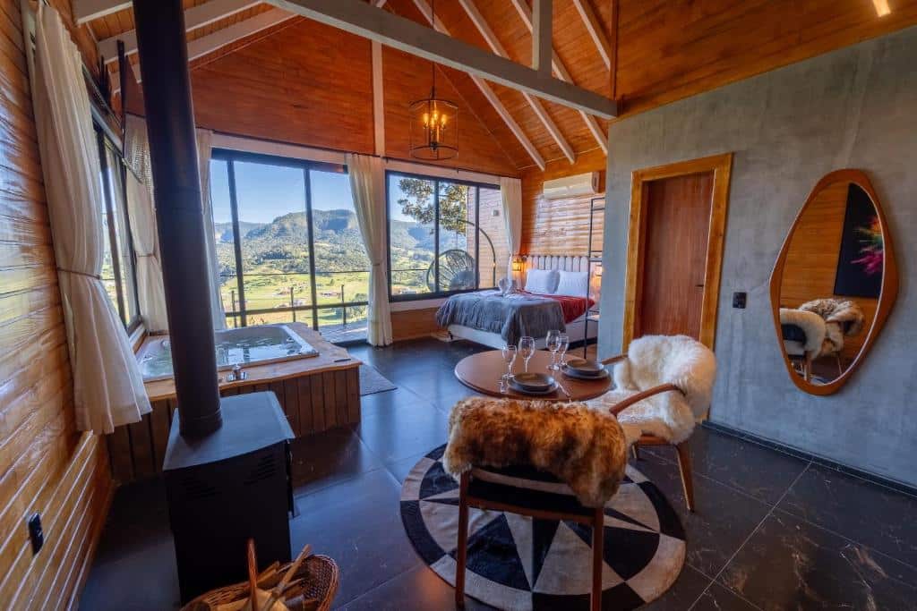 A imagem mostra o interior de um chalé no Hope Valley Chalés, decorado com um estilo rústico e moderno. O espaço é amplo, com teto alto e vigas expostas. Há uma cama de casal em frente a grandes janelas de vidro que proporcionam uma vista panorâmica das montanhas e da natureza ao redor. Próximo à janela, há uma banheira de hidromassagem de madeira. O ambiente também conta com uma lareira no centro, uma pequena mesa redonda com cadeiras cobertas por peles decorativas, além de detalhes de madeira nas paredes e uma atmosfera aconchegante.