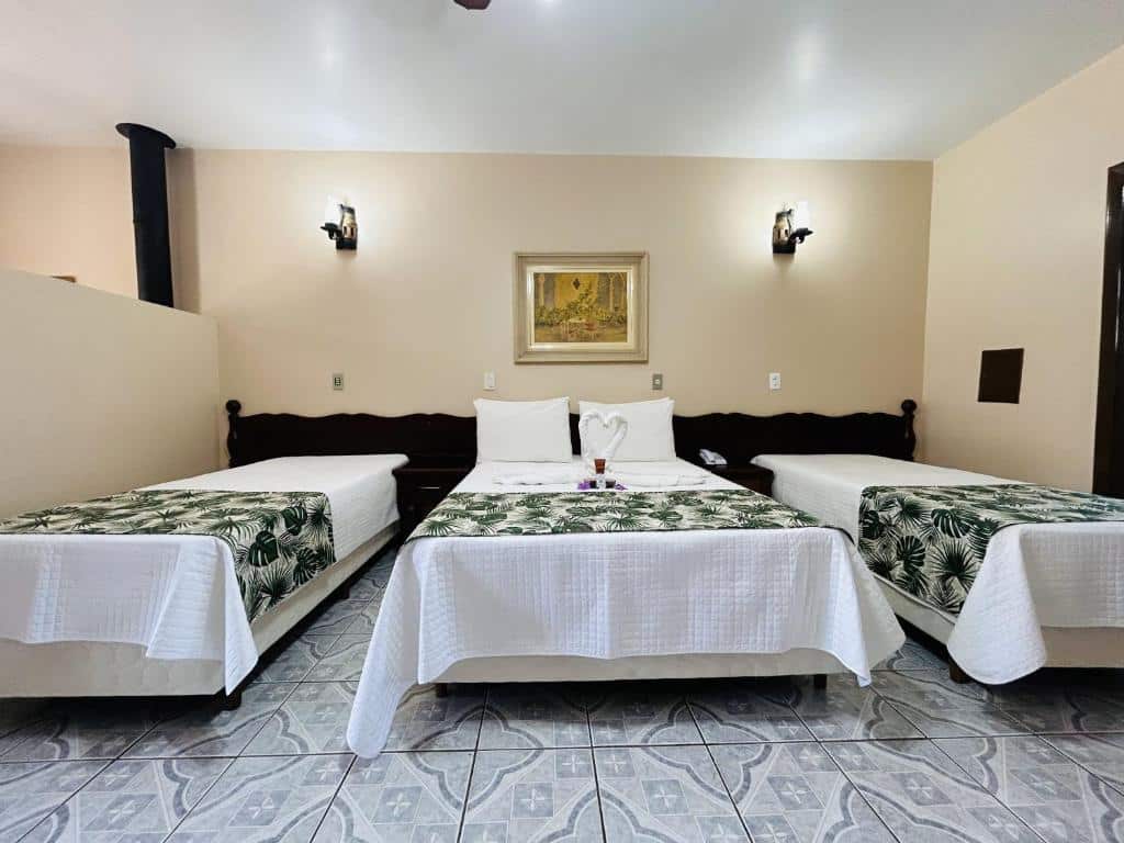 O quarto do hotel é espaçoso e decorado de forma simples e confortável. Há uma cama de casal no centro, com uma colcha branca, e duas camas de solteiro em cada lado, cobertas com mantas estampadas de folhas tropicais. Ao fundo, uma parede clara com uma pintura emoldurada dá um toque de elegância ao ambiente. O piso de azulejo possui desenhos geométricos, e a iluminação suave das arandelas na parede cria uma atmosfera acolhedora e relaxante, perfeita para descansar após um dia de atividades na fazenda.