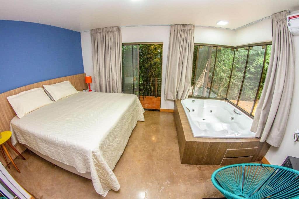 O quarto Hotel Fazenda Vale Alvorada no Paraná é espaçoso e moderno, com uma cama de casal grande ao lado de uma banheira de hidromassagem. As paredes em tons claros contrastam com um detalhe azul em uma das paredes, trazendo cor ao ambiente. Grandes janelas de vidro permitem a entrada de luz natural e oferecem uma vista para a vegetação externa, com cortinas claras que podem ser fechadas para privacidade. O ambiente é decorado de forma simples e aconchegante, com uma cadeira colorida de estilo contemporâneo e uma mesa de cabeceira ao lado da cama.