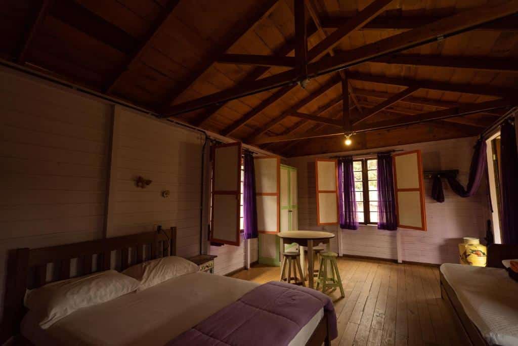 A imagem mostra o interior de um quarto na Pousada Aguaraguazu, com um estilo rústico e acolhedor. O ambiente é composto por paredes de madeira pintadas de branco e um teto alto com vigas expostas, também de madeira. O quarto é espaçoso e iluminado por duas janelas com cortinas roxas esvoaçantes, que deixam a luz natural entrar suavemente. Há duas camas simples com lençóis brancos e cobertores em tom de lilás. No centro do quarto, há uma pequena mesa redonda de madeira clara com dois banquinhos, criando um espaço aconchegante para refeições ou conversas.