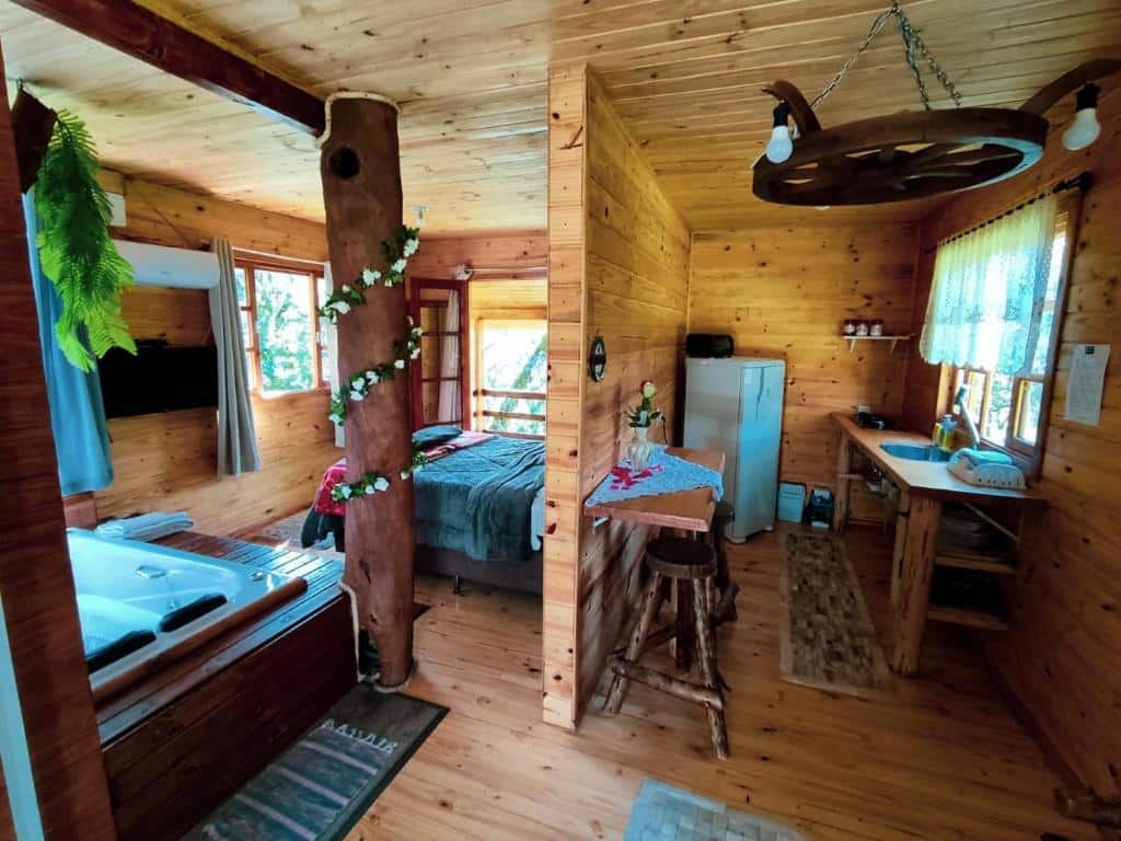 A imagem mostra o interior do Pousada Recanto do Lobo Chalés em Urubici com decoração rústica, predominantemente em madeira. No centro, há uma estrutura de tronco de árvore decorado com plantas. O ambiente é dividido entre uma pequena cozinha equipada com geladeira e pia, e uma área de descanso com cama e uma banheira de hidromassagem ao lado. O espaço é aconchegante, com janelas que proporcionam boa iluminação natural e uma atmosfera acolhedora.