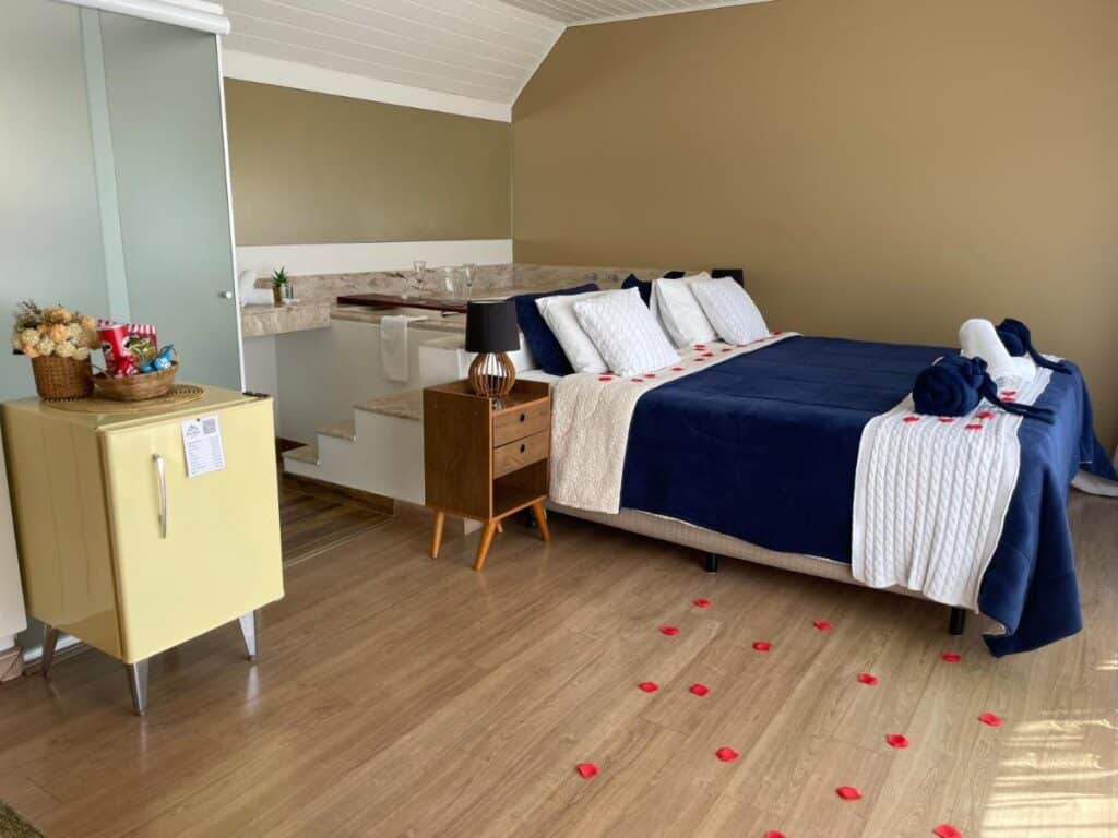 A imagem mostra um quarto aconchegante no Bela Vista Chalé com uma cama de casal grande, decorada com uma colcha azul escura e almofadas brancas. Pétalas de rosa vermelhas estão espalhadas pela cama e pelo chão, criando um clima romântico. Ao lado da cama, há um criado-mudo de madeira com um abajur preto. No canto do quarto, há uma pequena geladeira retrô amarela com uma cesta de lanches sobre ela. Ao fundo, uma banheira de hidromassagem está posicionada em um degrau elevado, com toalhas e copos dispostos ao lado, proporcionando um ambiente relaxante e acolhedor.
