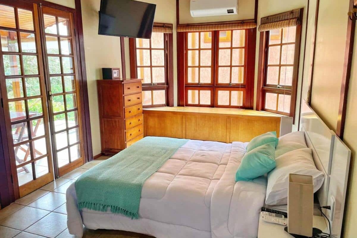 A imagem mostra um quarto bem iluminado com uma cama de casal coberta por um edredom branco e uma manta verde clara no Chalé das Águas. Há almofadas combinando em tons de verde e azul. À esquerda, portas de vidro dão acesso a uma varanda, permitindo a entrada de luz natural. Na parede oposta, há janelas amplas com molduras de madeira, um ar-condicionado e uma TV montada na parede. Um armário alto de madeira com gavetas está ao lado da cama, complementando o ambiente acolhedor e organizado. O piso é de cerâmica clara.