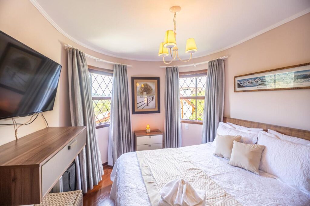 A imagem mostra um quarto aconchegante do Chalé dos Sonhos, com paredes claras e três janelas com molduras de madeira e cortinas cinzas. As janelas têm um design de grade, permitindo a entrada de luz natural. No centro do quarto, há uma cama grande com colcha branca e almofadas bege. Acima da cama, há um quadro de paisagem, e ao lado, um criado-mudo com uma pequena vela acesa. Uma televisão está fixada na parede em frente à cama, com um móvel de madeira logo abaixo. O ambiente é iluminado por um lustre simples com três lâmpadas.