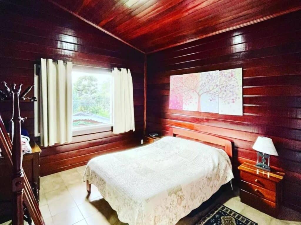 A imagem mostra um quarto aconchegante com paredes de madeira envernizada em tom escuro, criando uma atmosfera acolhedora no Chalé Manacá House. Ao centro, há uma cama de casal coberta com uma colcha clara e simples. À direita da cama, há uma mesa de cabeceira com um abajur branco pequeno, e na parede acima da cama, um quadro dividido em três partes com a imagem de uma árvore em cores pastéis. A janela grande ao fundo está parcialmente coberta por cortinas brancas, permitindo a entrada de luz natural. Ao lado esquerdo, há um cabideiro de madeira.