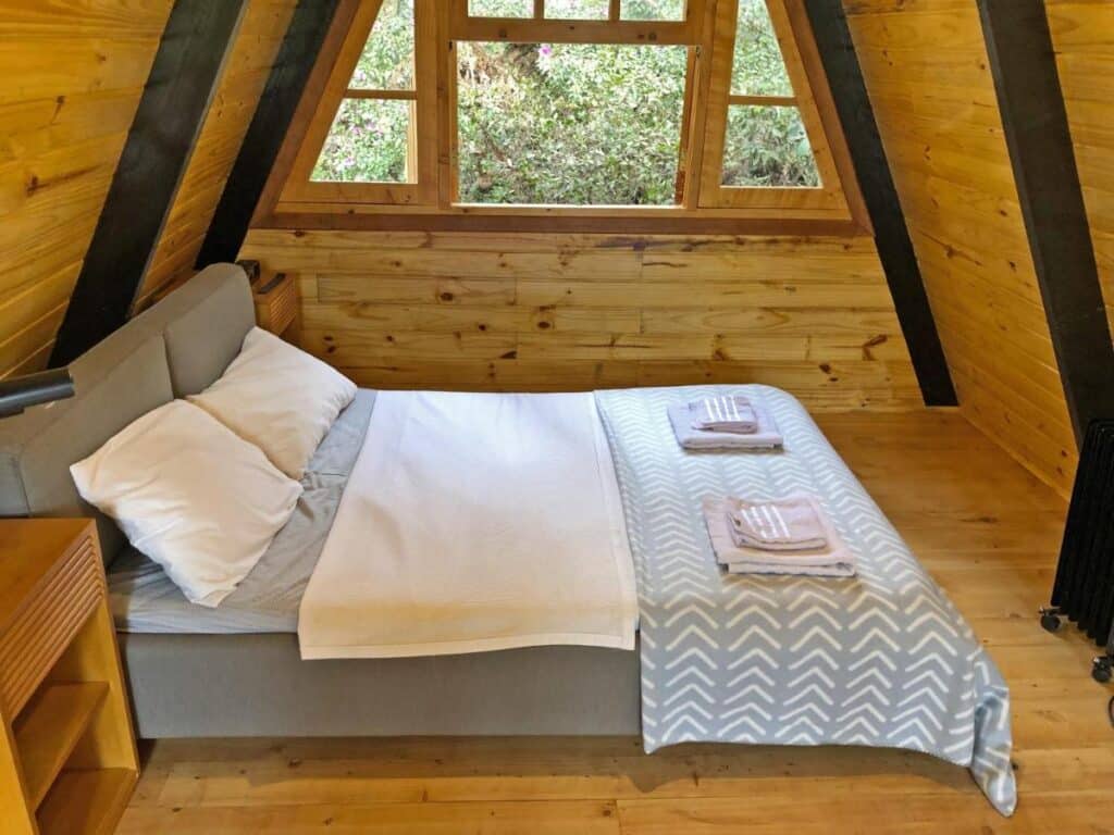 A imagem mostra o interior de um quarto do Chalé Mon Amour com paredes e teto revestidos de madeira clara. A cama de casal, com cabeceira acolchoada cinza, está coberta por um cobertor com estampa geométrica em tons de cinza e branco, e duas toalhas dobradas foram colocadas sobre ela. A grande janela triangular na parede de fundo oferece uma vista para a vegetação externa, trazendo luz natural ao ambiente. Ao lado da cama, há uma mesa de cabeceira de madeira simples, e o chão também é de madeira, criando uma sensação de aconchego e simplicidade.