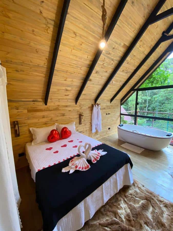 A imagem mostra um quarto romântico com decoração de madeira clara e teto inclinado com vigas pretas expostas. No centro, há uma cama de casal decorada com corações vermelhos e duas toalhas dobradas em formato de cisnes. Ao lado da cama, há uma banheira branca, posicionada próxima a uma grande janela de vidro que oferece vista para uma área verde do lado de fora. Dois roupões brancos estão pendurados na parede de madeira. O ambiente é iluminado por uma luz pendente simples, criando uma atmosfera aconchegante e íntima.