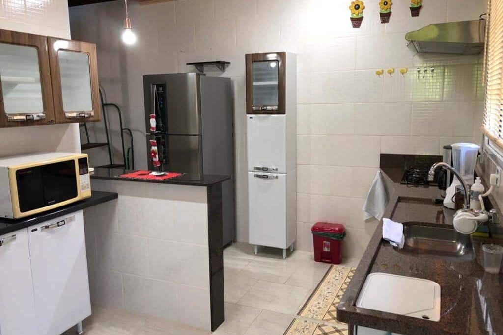 A imagem mostra uma cozinha organizada e moderna com armários brancos e detalhes em madeira nas portas superiores, no Chale Premium Casal e Chale Charming Casal. No centro, há uma pequena bancada preta com um micro-ondas em cima. Ao fundo, há uma geladeira prateada e um freezer branco ao lado. A pia tem uma bancada de granito escuro e está equipada com uma torneira moderna, próxima a um fogão embutido. Acima da pia, há uma janela com persianas que ilumina o ambiente. Decorações de flores amarelas enfeitam a parede clara, criando um toque alegre e acolhedor.