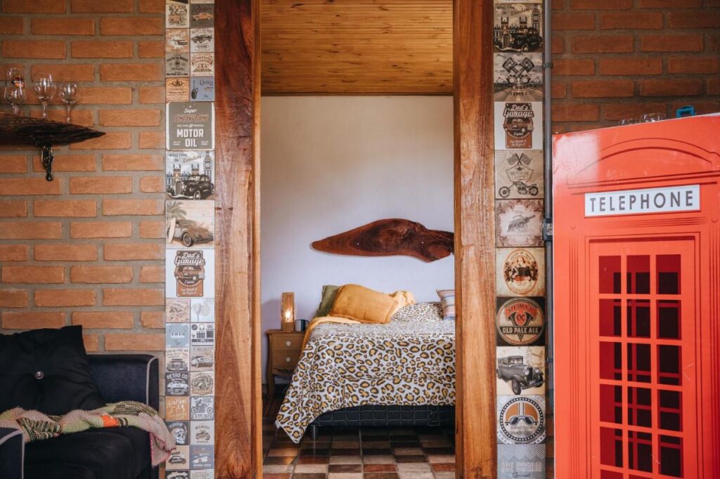 A imagem mostra o interior do Chalé Refugio da Pedra SP com uma decoração temática e criativa. À direita, há uma réplica de cabine telefônica vermelha ao estilo britânico. No centro, uma porta de madeira leva a um quarto com cama de estampa de oncinha e almofadas em tons de amarelo e laranja. As paredes ao redor da porta são decoradas com placas de estilo retrô, exibindo propagandas antigas de carros e motos. À esquerda, parte de um sofá preto com um cobertor colorido aparece na imagem, com uma prateleira de madeira acima exibindo taças de vidro.