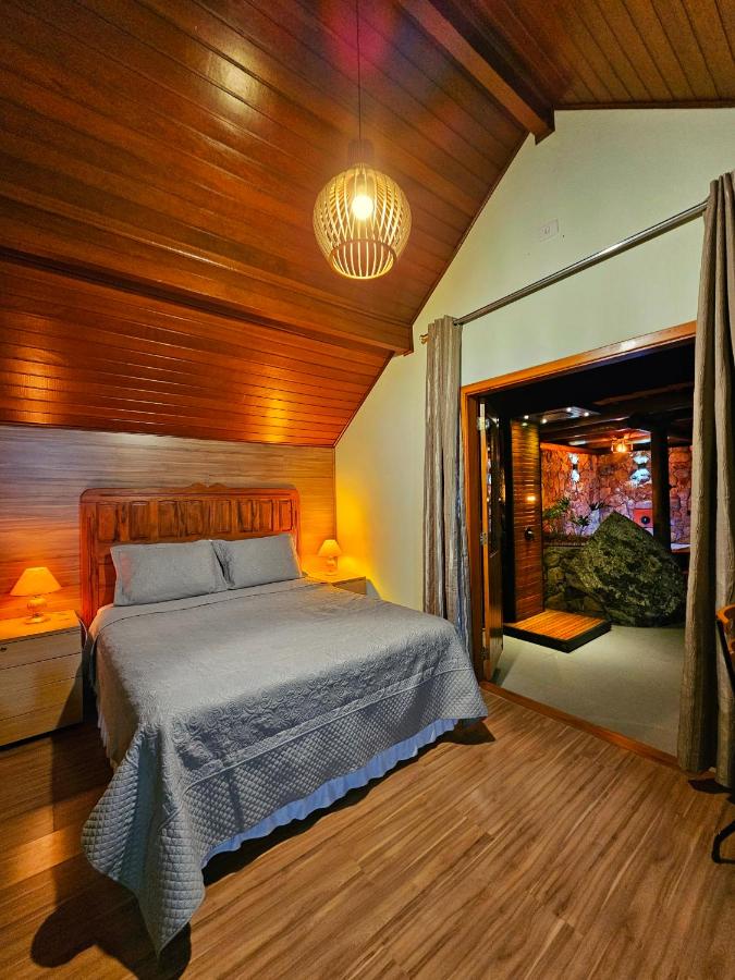 A imagem mostra um quarto aconchegante com teto de madeira inclinada, iluminado por uma luminária pendente de design rústico nos Chalés Araucária e Manacá. A cama de casal tem uma cabeceira de madeira trabalhada e está coberta com uma colcha cinza. Nas laterais da cama, há pequenos criados-mudos com luminárias acesas, criando uma iluminação suave. À direita, uma porta aberta revela uma área com pedras e iluminação indireta, adicionando um toque natural ao ambiente. O piso é de madeira clara, completando o estilo acolhedor do quarto.