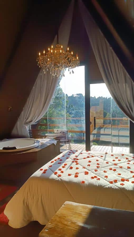 A imagem mostra o interior romântico de uma cabana com um quarto decorado para uma ocasião especial nos Chalés Capivari Diamante. A cama de casal está coberta por uma colcha branca e decorada com pétalas de rosas vermelhas. Ao lado da cama, há uma banheira de hidromassagem posicionada em um canto, com vista para o exterior através de uma porta de vidro que dá para uma varanda. O ambiente é iluminado por um lustre de cristal pendurado no teto, que adiciona um toque elegante ao espaço. Ao fundo, pode-se ver uma bela paisagem natural com árvores ao longe.