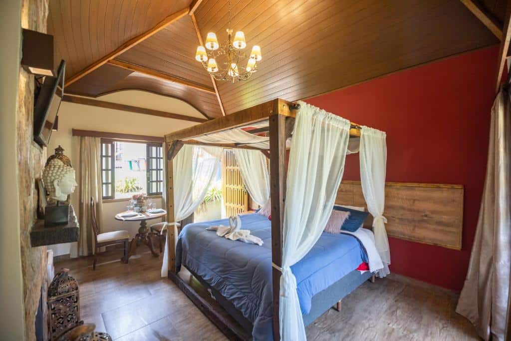 A imagem mostra o interior de um quarto dos Chalés da Mantiqueira com uma cama de dossel de madeira, coberta por lençóis azuis e com cortinas brancas esvoaçantes penduradas nas laterais. O teto de madeira tem vigas expostas, e um lustre pendurado no centro adiciona charme ao ambiente. A parede ao fundo é vermelha, e ao lado há uma mesa redonda com duas cadeiras próximas a uma janela, que permite a entrada de luz natural. No canto esquerdo, uma prateleira exibe uma pequena estátua decorativa de um Buda.