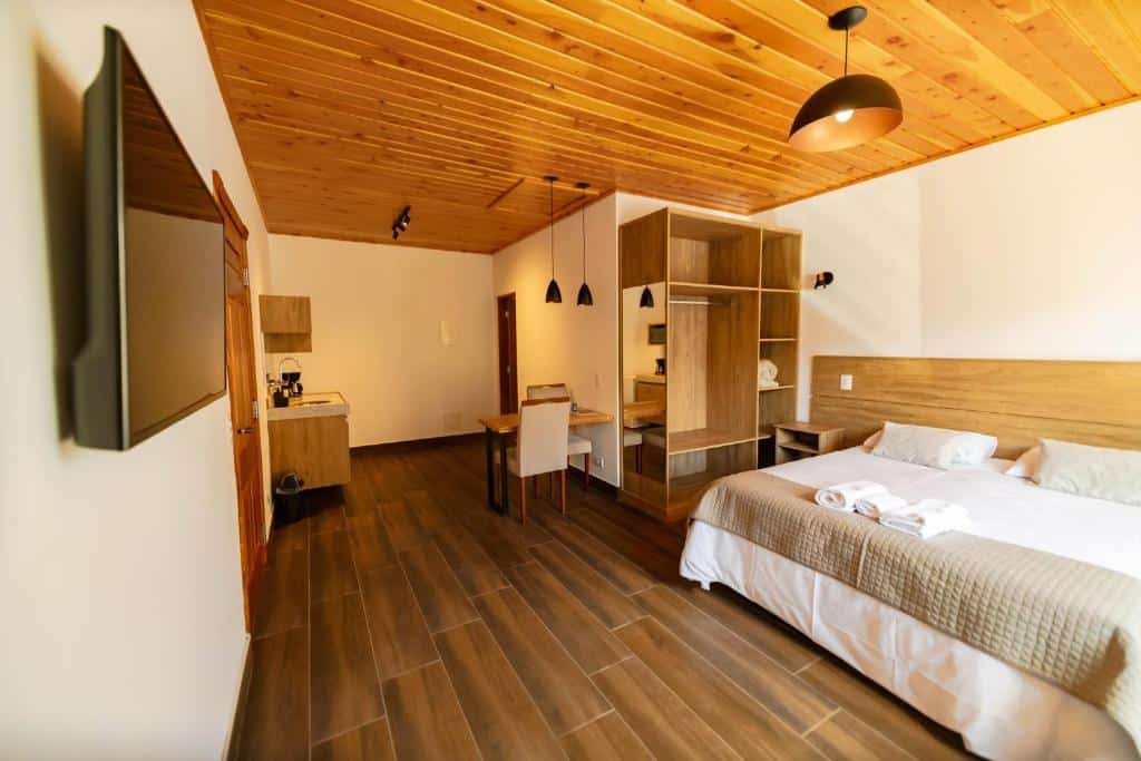 A imagem mostra um quarto espaçoso e moderno dos Chalés Estilo de Campos, com piso de madeira e teto de forro de madeira clara. À direita, há uma cama de casal grande, coberta com lençóis brancos e um edredom bege, com toalhas cuidadosamente dobradas sobre a cama. Na parede à esquerda, há uma televisão montada. O quarto possui uma mesa de trabalho com cadeira, prateleiras abertas para armazenamento e luminárias pendentes pretas. Ao fundo, há uma pequena área com uma cafeteira e itens de cozinha. O ambiente é bem iluminado, acolhedor e funcional.