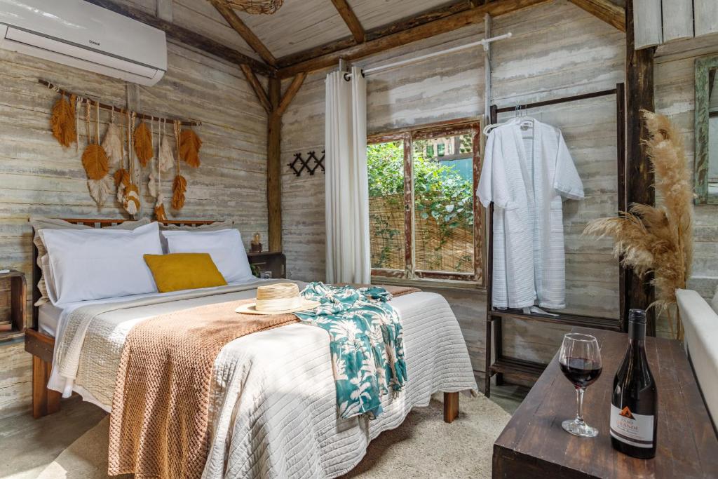 Quarto do Chalés Magia do Campeche com cama de casal do lado esquerdo da imagem e em frente a cama uma cômoda com uma taça e uma garrafa de vinho em cima. Representa chalés em Florianópolis.