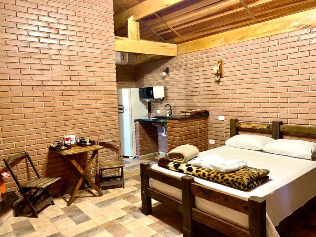 A imagem mostra um quarto rústico com paredes de tijolos aparentes e móveis de madeira nos Chalés Marinelli. No centro, há uma cama de casal com lençóis brancos e cobertores dobrados, incluindo um de estampa de oncinha. Ao lado da cama, há uma pequena mesa de madeira com duas cadeiras, onde estão colocados alguns itens como xícaras. Ao fundo, há uma cozinha compacta com pia, micro-ondas e geladeira branca. O teto é de madeira com vigas expostas, dando um toque acolhedor e rústico ao ambiente, com iluminação suave. O piso é composto por ladrilhos em tons terrosos.