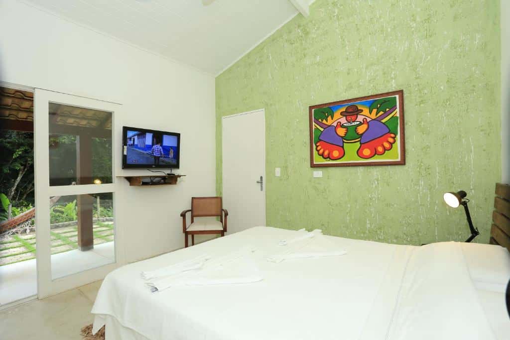 A imagem mostra um quarto claro e moderno com uma cama de casal coberta com lençóis brancos. A parede à direita é pintada de verde claro com textura, decorada com um quadro colorido e vibrante, representando uma figura estilizada. À frente da cama, há uma televisão presa à parede, acima de uma pequena prateleira com objetos. Ao lado, uma cadeira de madeira com estofado claro está posicionada perto de uma porta de vidro, que leva a uma varanda externa com rede, permitindo a entrada de luz natural e uma vista para a vegetação. Os Chalés Mariolinha fazem parte da lista de chalés para casal no interior de São Paulo.