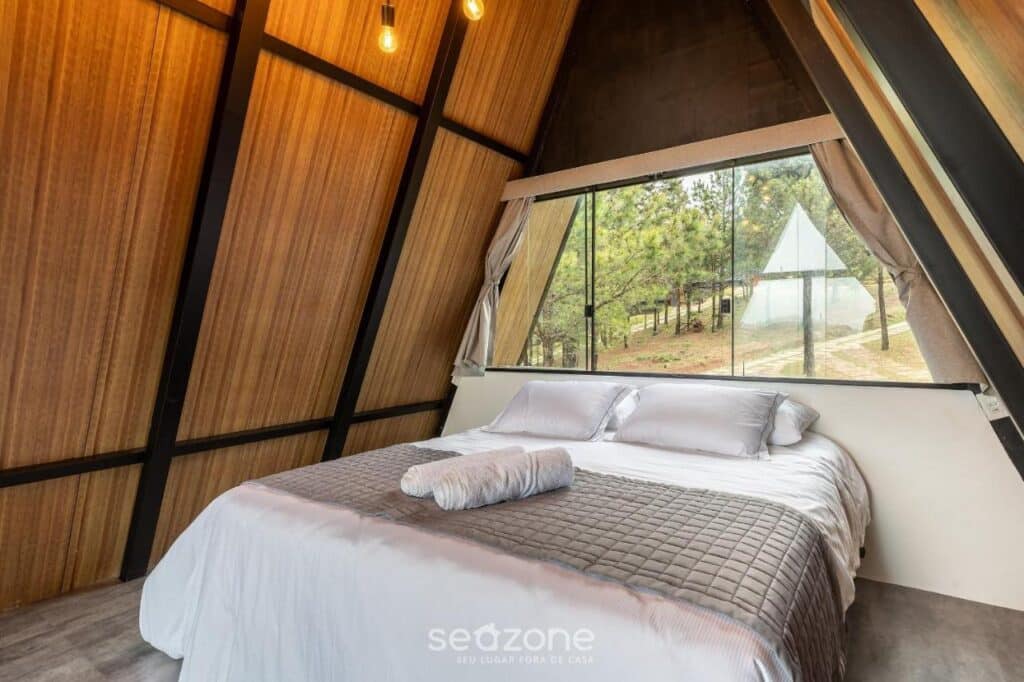A imagem mostra um quarto com design moderno e minimalista em formato triangular nos Chales Naturale. A cama de casal ocupa a maior parte do espaço, com roupa de cama clara e uma manta cinza sobreposta. As paredes são revestidas de madeira e, à direita da cama, há uma ampla janela de vidro que oferece uma vista para uma área arborizada, permitindo a entrada de muita luz natural. As cortinas estão abertas, e há uma lâmpada pendente no teto, reforçando o estilo simples e aconchegante do ambiente. Esse local é uma recomendação entre os chalés em Campos do Jordão.