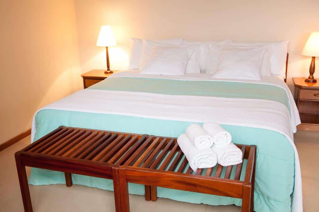 Quarto do Hotel Estancia Betania com cama de casal no centro do quarto com uma cômoda em cada lado da cama com luminária e no pé da cama um banco de madeira. Representa dos hotéis fazenda no Paraná.