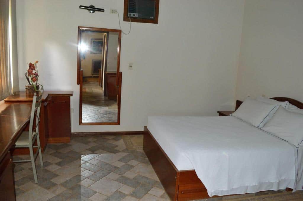 Quarto do Hotel Lago das Pedras com cama de casal do lado direito da imagem e em frente a cama uma mesa com cadeira. Representa dos hotéis fazenda no Paraná.