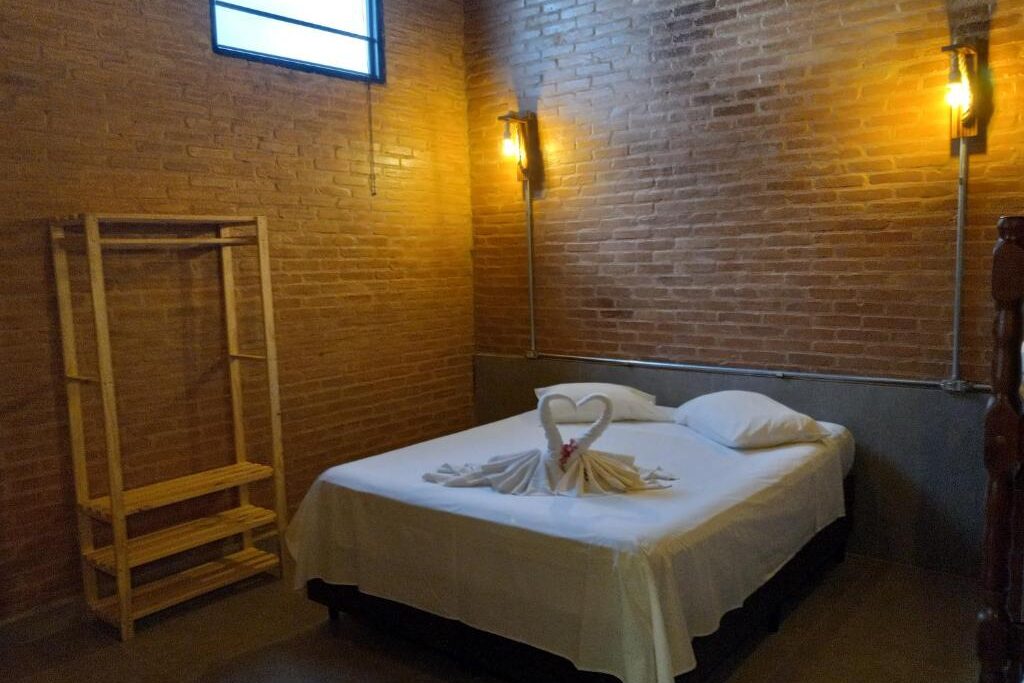 A imagem mostra um quarto simples e acolhedor com paredes de tijolos aparentes e iluminação suave de arandelas nas laterais da cama, na La Villa Chalés. A cama de casal está coberta com um lençol branco e decorada com toalhas dobradas em formato de cisnes, criando um toque romântico. Há uma pequena janela no alto da parede, deixando entrar luz natural. No canto esquerdo, há um cabideiro de madeira simples, sem portas. O ambiente é rústico, com uma decoração minimalista e aconchegante.