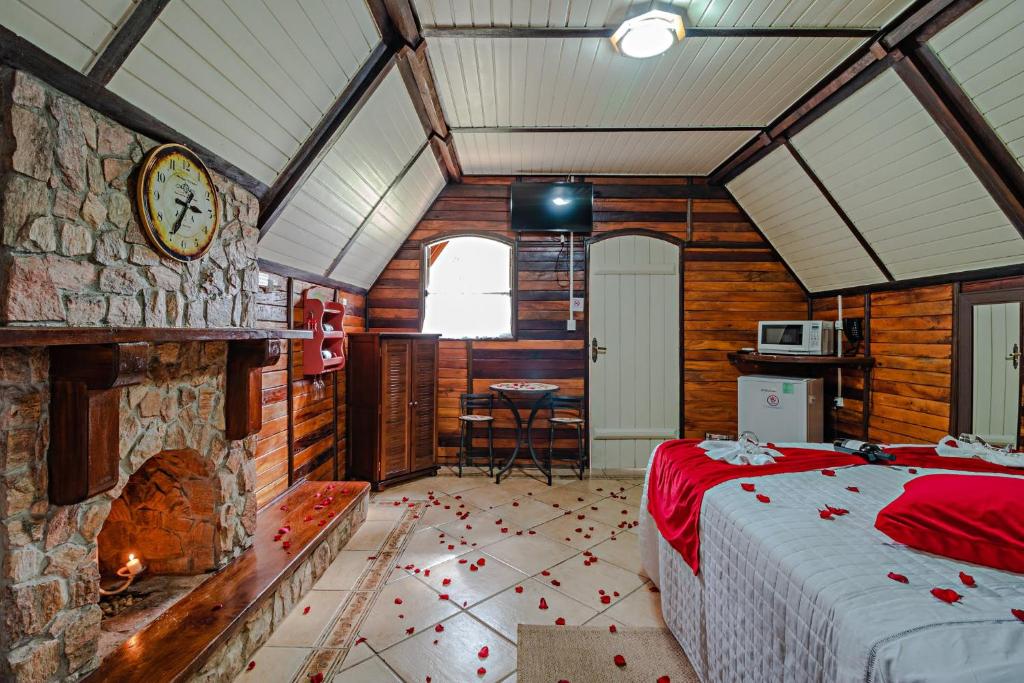 A imagem mostra o interior de uma cabana de madeira com paredes de tijolos e vigas de madeira escura no Mirágua Refugios Pousada. Há uma lareira de pedra à esquerda, com um relógio grande acima. Sobre a cama de casal, coberta por uma colcha branca com detalhes em vermelho, pétalas de rosas estão espalhadas pelo chão e pela cama. No fundo, há uma pequena mesa com cadeiras, um armário de madeira, um micro-ondas e um frigobar. O ambiente tem uma iluminação aconchegante e rústica, com um clima romântico.