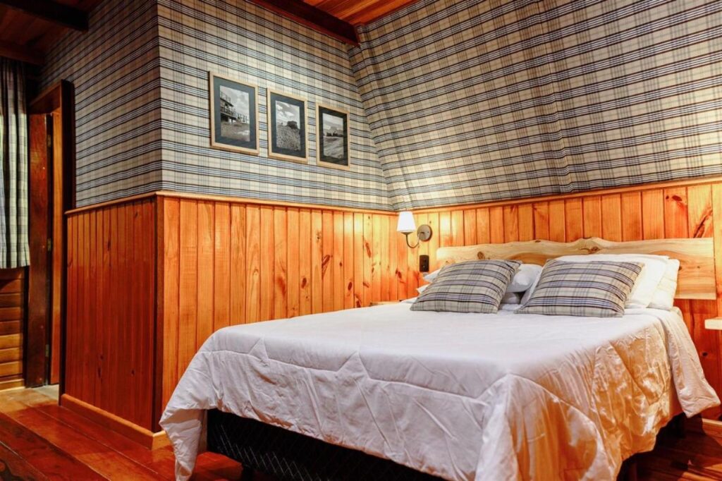 A imagem mostra o interior de um quarto aconchegante da Pousada Chalés Por do Sol, com paredes revestidas de madeira até a metade e um papel de parede xadrez nas partes superiores. A cama de casal está coberta por uma colcha branca e tem duas almofadas com estampa combinando com o padrão xadrez da parede. Acima da cama, há três quadros com fotos em preto e branco, e uma luminária de parede ao lado, criando um ambiente confortável e acolhedor, com um estilo rústico. O piso também é de madeira, completando a atmosfera de chalé.