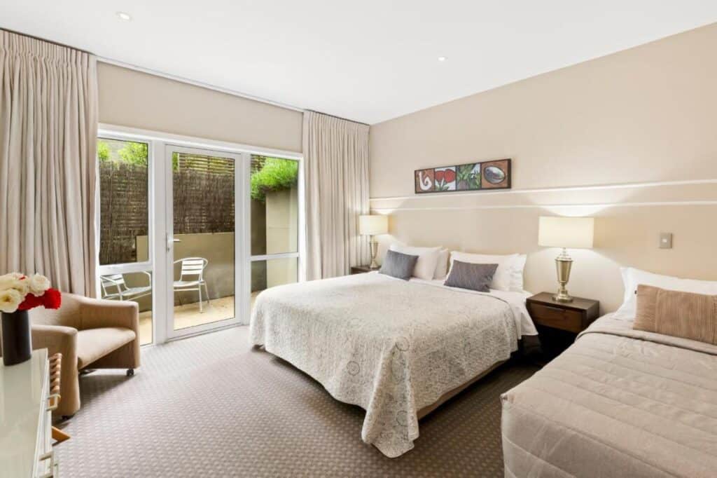 Um quarto do B&B Queenstown House Bed & Breakfast and Apartments. Ele tem duas camas bem arrumadas, uma pequena mesa e cadeiras no fundo, uma cadeira à esquerda e uma porta com janela que dá para fora.