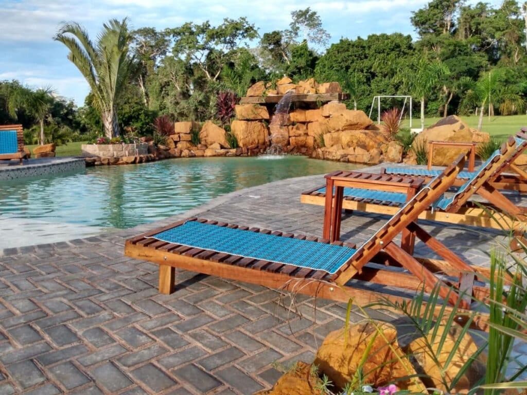 Área externa do Recanto da Natureza. Cadeiras de tomar sol do lado direito, do lado esquerdo uma piscina com cascata, no fundo o gramado e árvores.