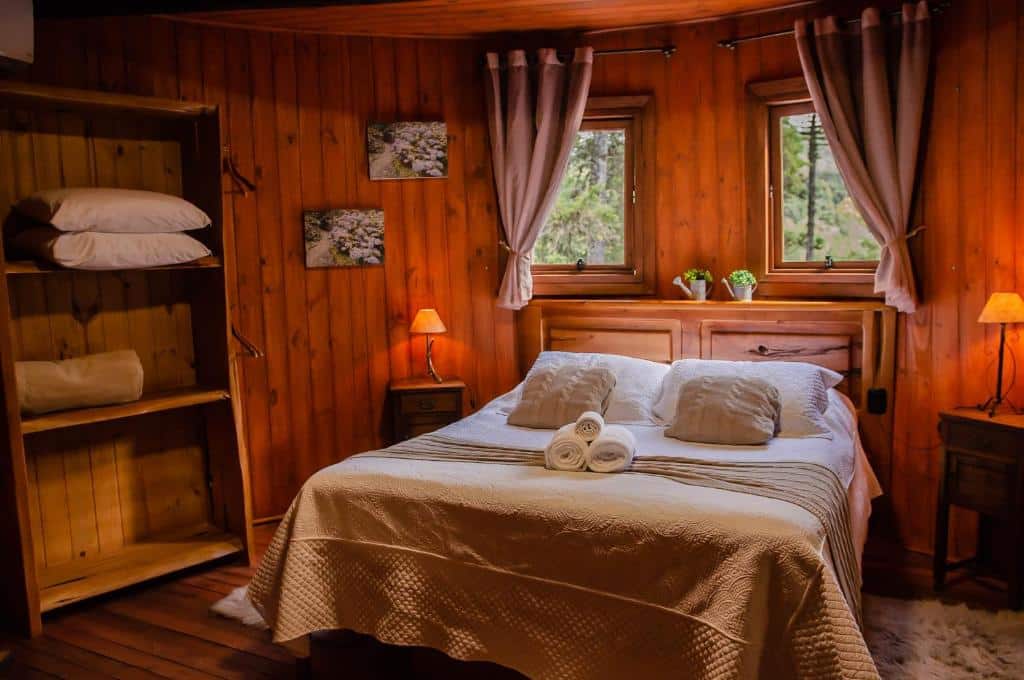 Quarto do Refúgio do Caracol Pipas e Chalés. As paredes, o chão e o teto são todos de madeira, criando uma atmosfera quente e rústica. No centro do quarto, há uma cama de casal com roupa de cama branca e duas toalhas enroladas aos pés da cama.
Ao lado da cama, há mesas de cabeceira com abajures combinando. Acima da cabeceira, há uma janela que permite a entrada de luz natural, ladeada por cortinas. Em frente à cama, há uma estante aberta com cobertores ou toalhas dobradas e itens decorativos.
Nas paredes, há quadros ou obras de arte que complementam a decoração. No chão, ao lado da cama, há um pequeno tapete, adicionando um toque extra de conforto ao ambiente.