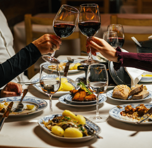 Imagem de uma mesa da Adega São Nicolau com vários pratos com pratos dos estabelecimentos taças de vinhos.