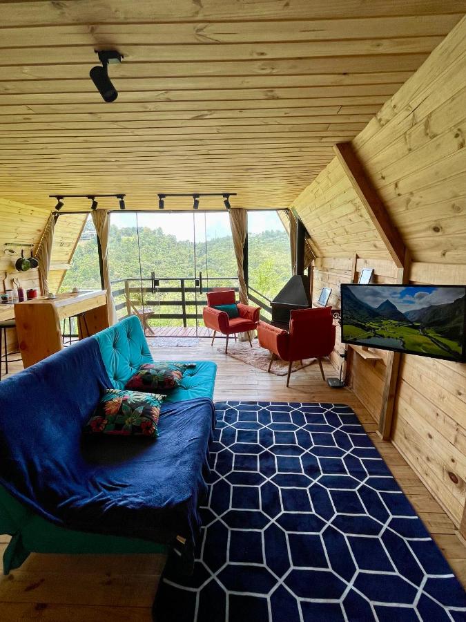 A imagem mostra o interior aconchegante de uma cabana de madeira com decoração moderna e rústica nos Chalés entre Nuvens. O espaço possui um sofá azul com almofadas coloridas, um tapete azul escuro com padrões geométricos, e duas poltronas vermelhas posicionadas perto de uma lareira triangular preta. Uma televisão está fixada na parede à direita, exibindo uma paisagem de montanha. Ao fundo, uma grande janela de vidro oferece uma vista panorâmica da floresta e montanhas, permitindo a entrada de muita luz natural. Uma pequena varanda está visível do lado de fora, acessada por uma porta de vidro.