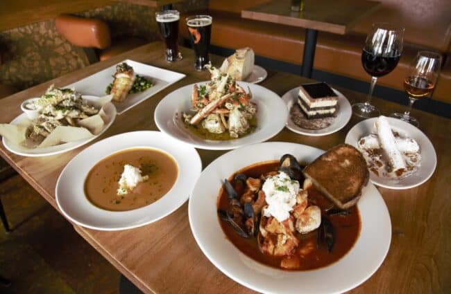 Uma mesa com vários pratos, incluindo sopa, frutos do mar, pão, sobremesa, duas taças de vinho e duas taças de cerveja preta no Scoma's Of Sausalito.