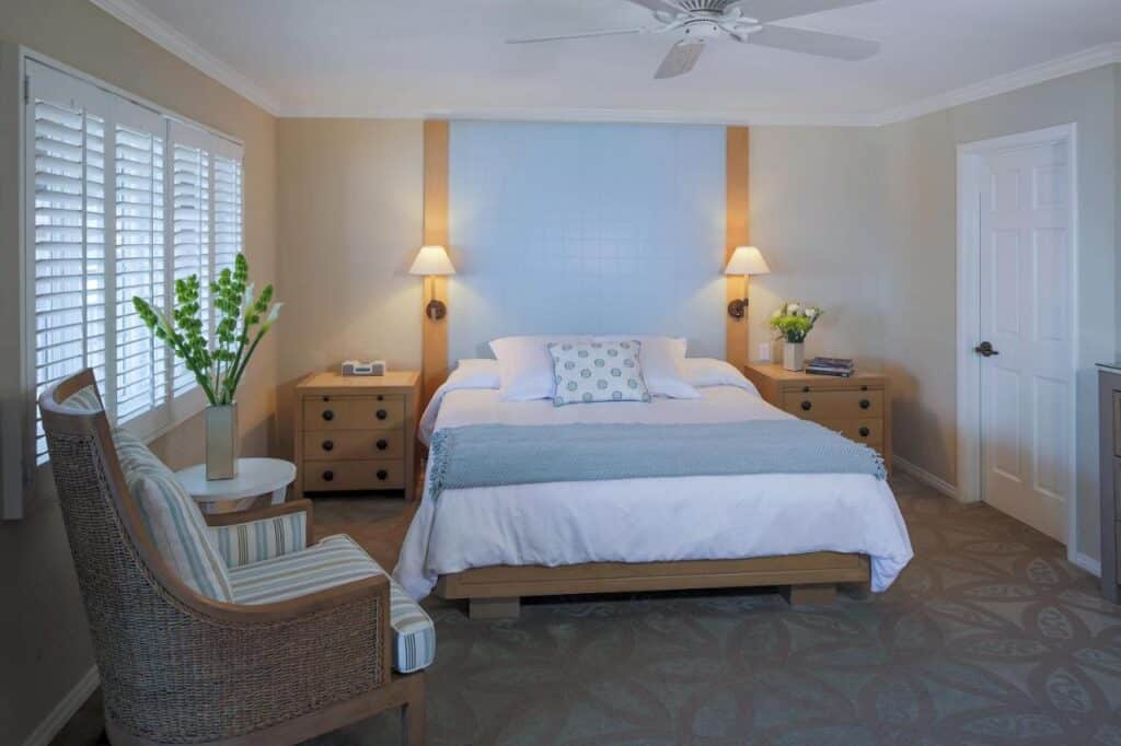 Quarto do Scripps Inn, com uma cama de casal e uma poltrona do lado esquerdo. Uma janela grande e uma mesinha com um vaso de flor também estão desse lado.