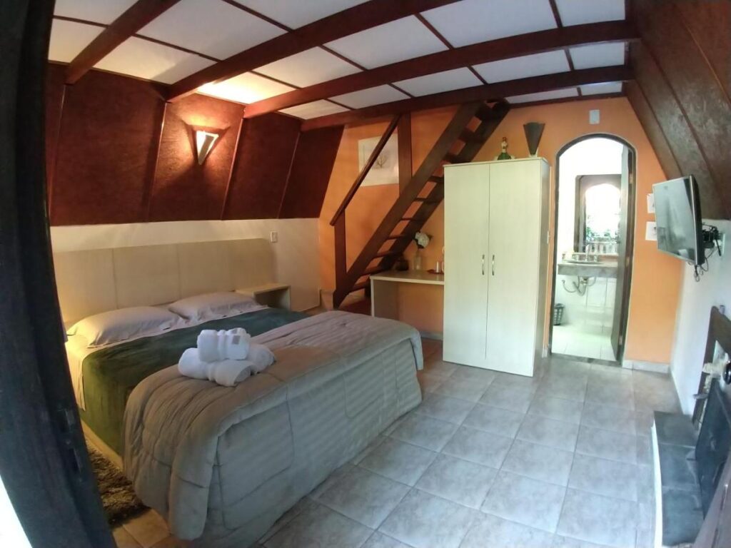 Quarto do Spazio di Garda, um dos chalés em São Roque. Uma cama de casal do lado esquerdo, do lado direito uma TV. No fundo uma escada, um guarda-roupa e a porta do banheiro.