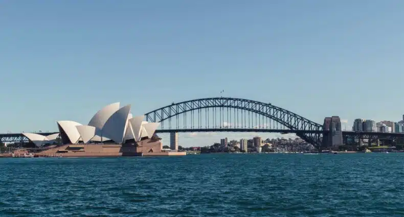 Vista da Ópera de Sydney e da Ponte da Baía de Sydney através da água sob um céu azul claro. A foto ilustra o post de o que fazer em Sydney.