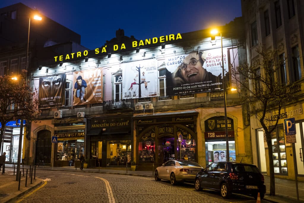 Imagem da frente do Teatro Sá da Bandeira com cena de rua à noite com a fachada iluminada do Teatro Sá da Bandeira, no Porto com cartazes e carros estacionados na frente. Representa o que fazer à noite no Porto.