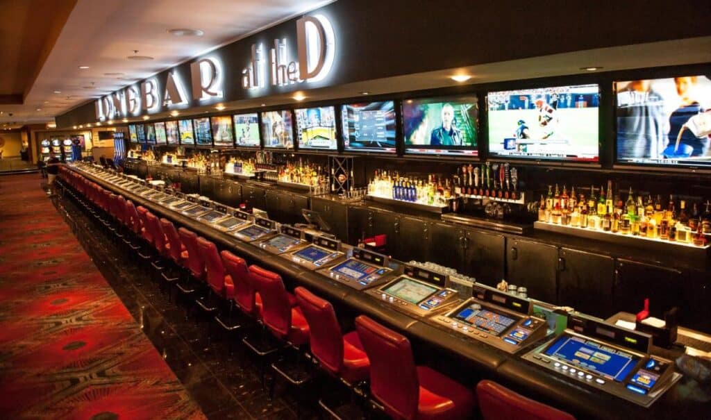 Vista do Sport Book no The D Las Vegas. Se trata de um bar de cassino com uma longa fileira de máquinas de vídeo pôquer, várias telas exibindo esportes e uma área de bar bem abastecida.