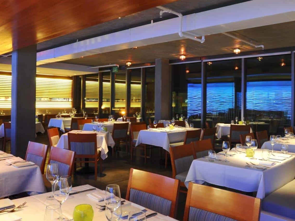 Restaurante no Tower 23 Hotel . Várias mesas estão no ambiente. Ao fundo há uma projeção de ondas do mar. Foto para o post de onde ficar em San Diego.