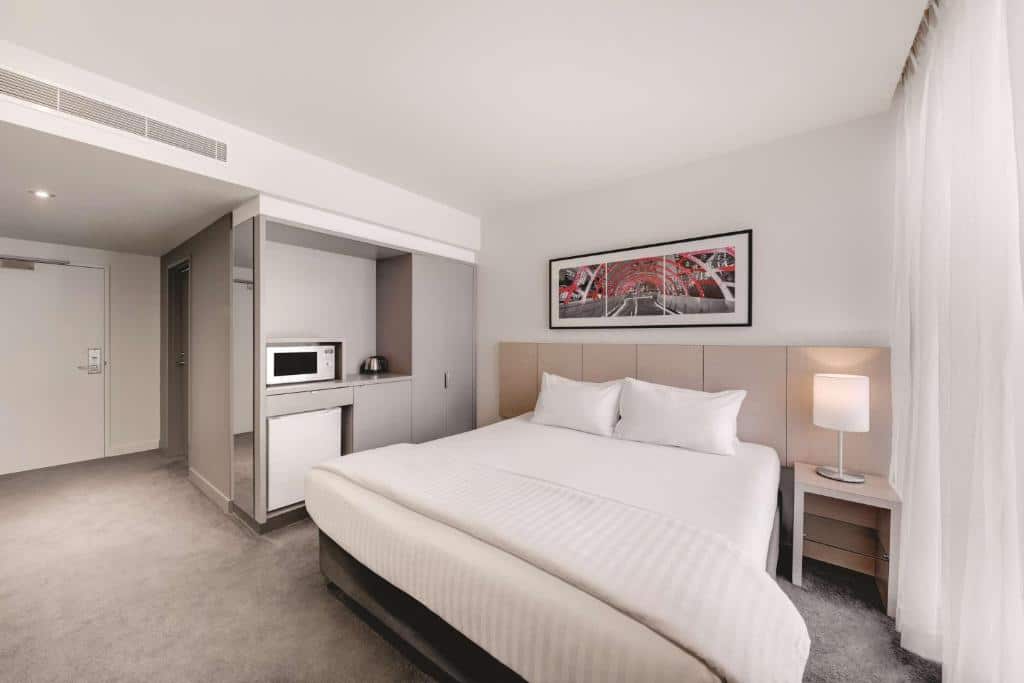 Um quarto do Travelodge Hotel Melbourne Docklands com uma cama grande, um abajur, um micro-ondas e um quadro emoldurado na parede, com cores neutras.