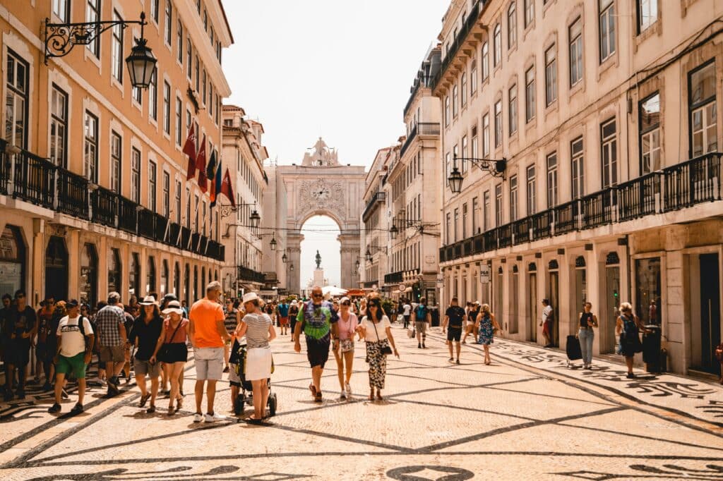Pessoas caminhando pelas ruas durante o dia com prédios antigos em cada lado da imagem, pessoas caminhando no centro e ao fundo um arco de concreto. Representa o que fazer em Lisboa.