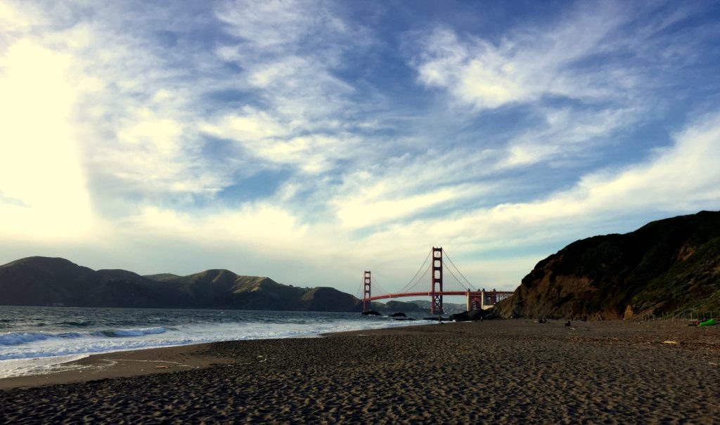 Baker Beach a com ondas suaves na costa, a Ponte Golden Gate ao fundo e um céu parcialmente nublado acima.