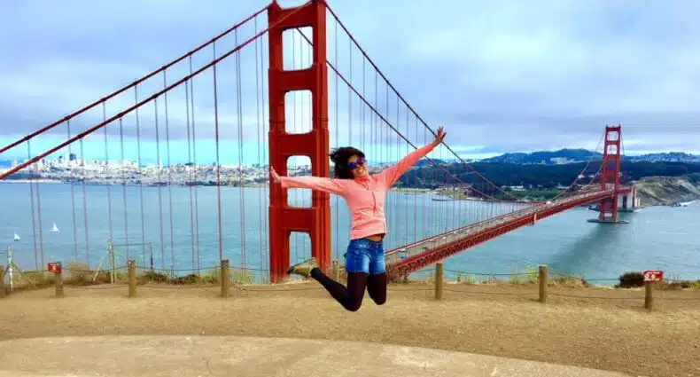 Pessoa pulando em frente à Ponte Golden Gate, vestindo um moletom rosa e óculos de sol, com água e paisagem urbana ao fundo. Imagem para ilustrar post sobre eSIM San Francisco.