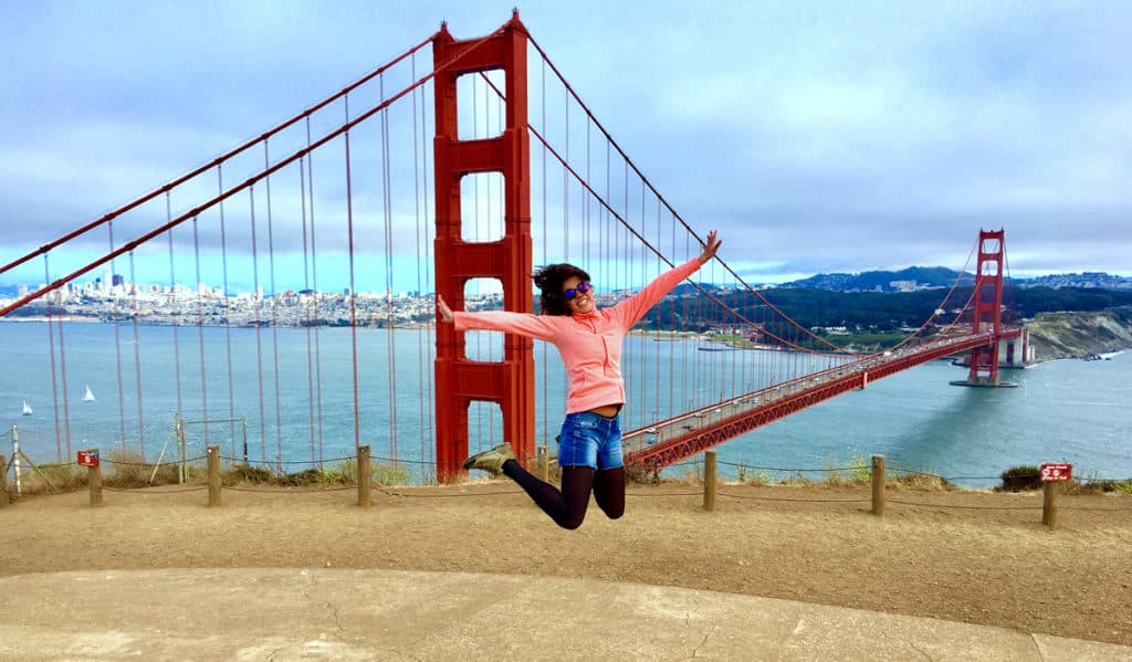 Pessoa pulando em frente à Ponte Golden Gate, vestindo um moletom rosa e óculos de sol, com água e paisagem urbana ao fundo.