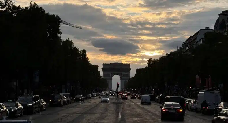Vista de uma rua movimentada da cidade que leva ao Arco do Triunfo, um dos passeios em Paris, sob um céu nublado ao pôr do sol, ladeada por árvores e carros em ambos os lados.