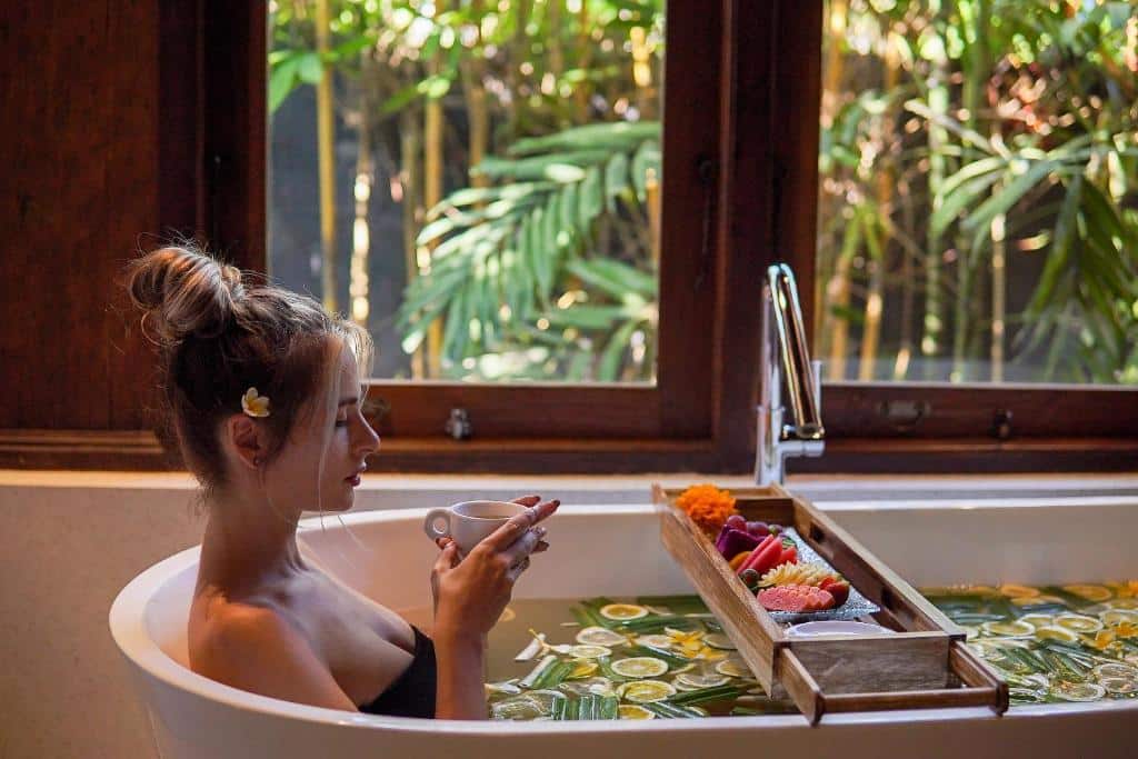 Uma mulher relaxa em uma banheira cheia de pétalas de flores e fatias de frutas cítricas, segurando uma xícara, com vista para uma vegetação exuberante do lado de fora da janela do Ubud Valley Boutique Resort.